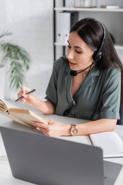 Teacher in headset reading book during online lecture on laptop in school 