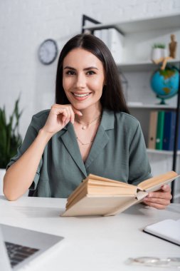 Smiling teacher holding book and looking at camera near blurred laptop in school 