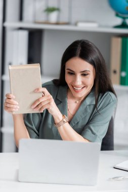 Smiling teacher holding book during online lesson on blurred laptop in school 