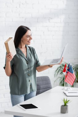 Sınıfta bayrakların yanında kitap ve dizüstü bilgisayar tutan neşeli öğretmen ve dijital tablet.