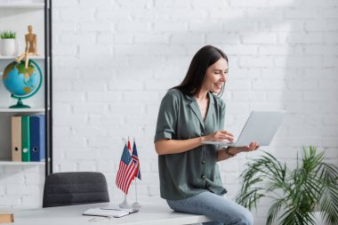 Cheerful teacher using laptop during online lecture near flags and notebook on table in school 