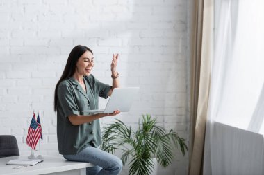 Cheerful teacher holding laptop during online lesson near flags on table in classroom 