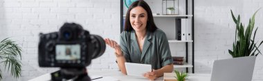 Positive teacher with digital tablet talking near blurred camera in school, banner 