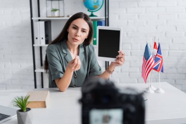 Teacher holding digital tablet and talking near camera and flags in classroom 