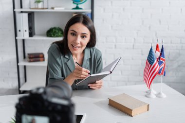 Smiling teacher holding notebook near book and blurred digital camera in class 