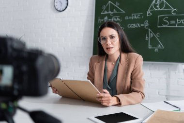 Teacher holding notebook near digital tablet and camera in classroom 