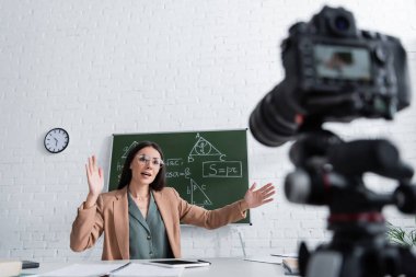 Teacher gesturing near chalkboard and blurred digital camera in school 