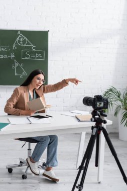 Teacher holding book while talking near digital camera in school 
