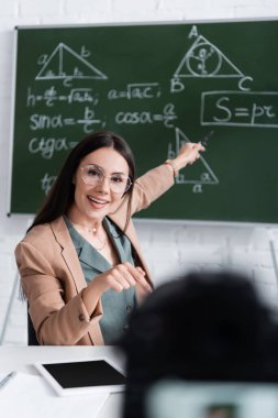 Okulda dijital kameranın yanında matematik formülleriyle bulanık tahtayı işaret eden pozitif öğretmen 