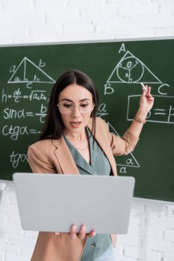 Teacher having video call on laptop near chalkboard with math formulas 