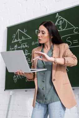 Teacher holding chalk and laptop during online lecture near chalkboard in classroom 