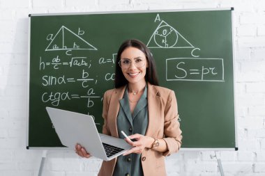 Matematik formüllerinin yanında tebeşir ve dizüstü bilgisayar tutan gülümseyen öğretmen. 