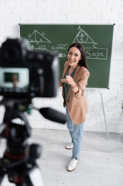 Positive teacher pointing at blurred digital camera and chalkboard in classroom 