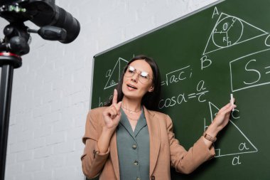 Teacher in eyeglasses explaining math formulas on chalkboard near digital camera in class 