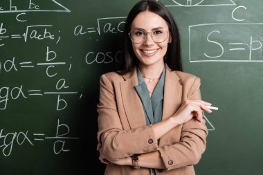 Positive teacher holding chalk near math formulas on chalkboard 