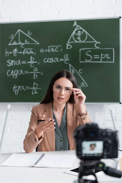 Teacher in eyeglasses talking near blurred digital camera and chalkboard