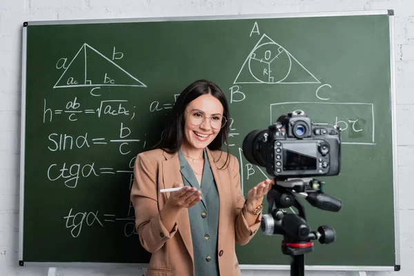 Positive teacher pointing at digital camera near chalkboard with math formulas