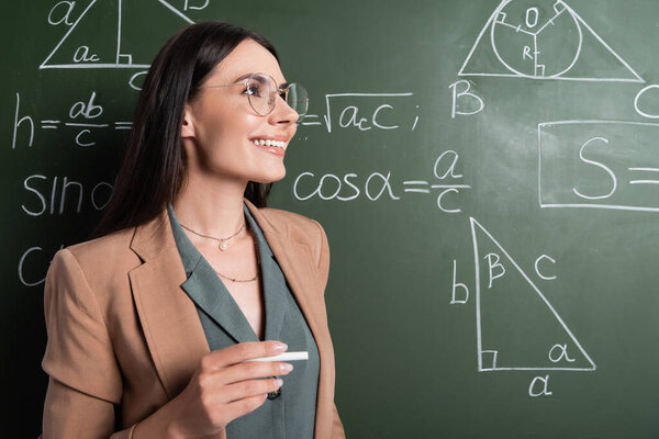 Happy teacher holding chalk near mathematic formulas on chalkboard 