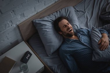Top view of man with insomnia yawning near clock and water on bedside table in morning