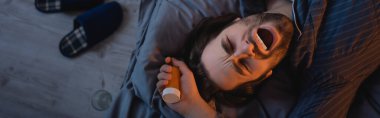 Top view of bearded man yawning and holding pills on bed at night, banner 