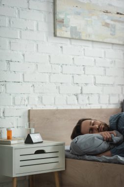 Young man with closed eyes lying on pillow near pills and alarm clock in bedroom 