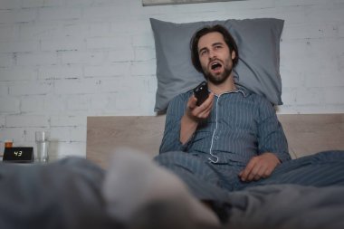 Man with sleep disorder yawning and holding remote controller while sitting on bed 