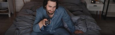 Pijama giymiş tatminsiz bir adam gece yatakta otururken kanalları tıklatıyor. 