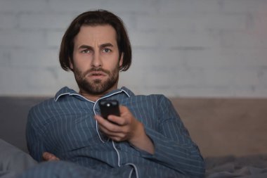 Displeased man in pajama watching tv while sitting on bed at night 