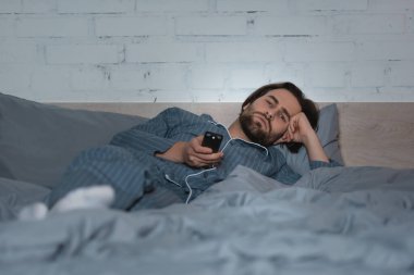 Yatakta yatarken elinde uzaktan kumandayla pijamalı yorgun bir adam. 