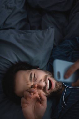 Top view of man with insomnia yawning and using smartphone on bed 