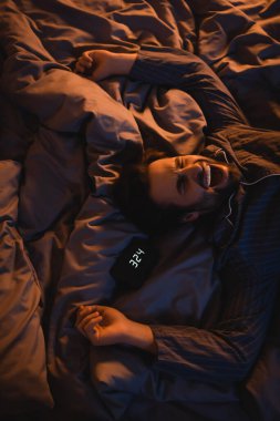 Top view of man in pajama yawning near alarm clock on bed at night 