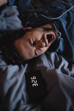 Top view of sleepless man yawning near alarm clock on bed at night 
