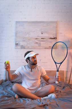 Confused sleepwalker in sportswear holding tennis ball and rocket on bed  clipart
