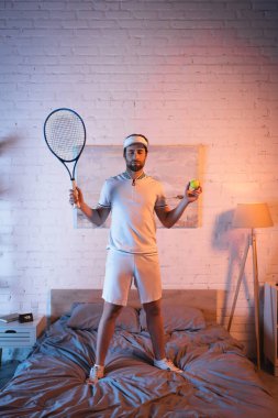 Sleepwalker in sportswear holding tennis rocket and ball on bed at night 