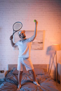 Somnambulist holding tennis ball and rocket while standing on bed at home 