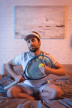 Noctambulist in sportswear holding tennis ball and rocket on bed at night 