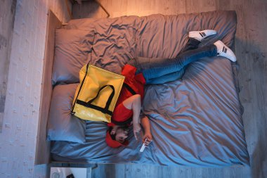 Top view of sleepwalker in delivery uniform pointing at smartphone on bed at night 
