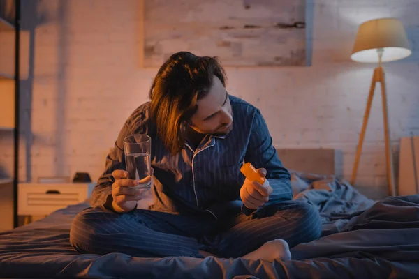 Man in pajama holding pills and water on bed at night