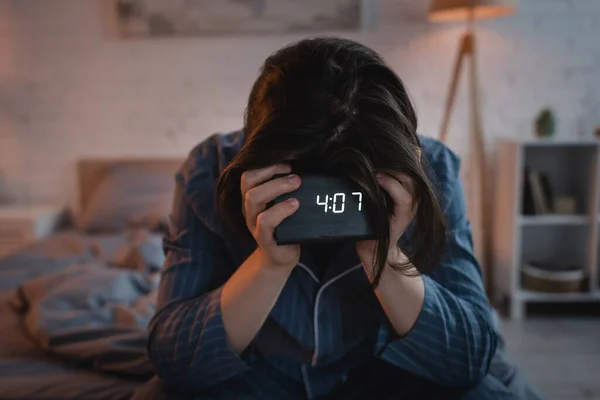 Young man with sleep disorder holding electronic clock on blurred bed at night