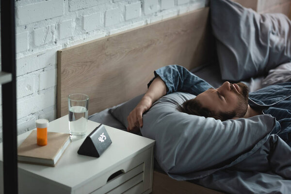 Мужчина, страдающий расстройством сна рядом с таблетками и будильником в спальне утром 