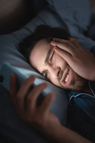 Tensed man with insomnia using blurred smartphone on bed at night