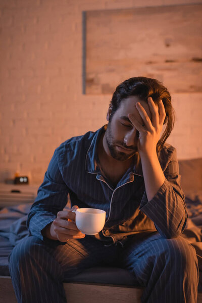 Exhausted young man in pajama holding cup while sitting on bed at night 