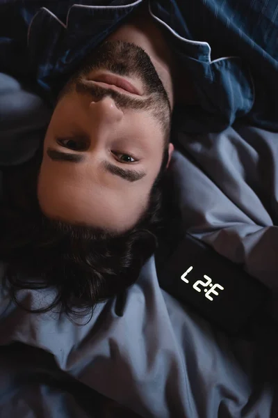 Top view of young man in pajama looking at camera near alarm clock on bed