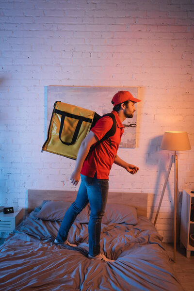 Side view of sleepwalker in uniform with thermo backpack standing on bed at night 