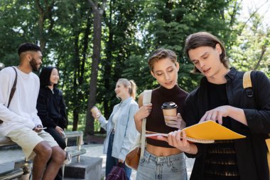 Student holding notebooks near friend with coffee to go in park 