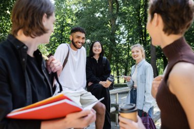 Cheerful multicultural students with smartphone and takeaway drink looking at blurred friends in park 