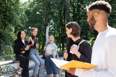 Excited multiethnic students holding coffee to go near blurred friends in park 