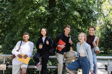 Multicultural students with notebooks and backpacks looking at camera in park 