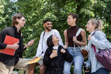 Smiling multicultural students with notebooks and smartphones talking in park 