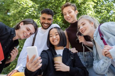 Asyalı bir öğrenci elinde kahve ile parktaki ırklar arası arkadaşlarıyla selfie çekiyor. 